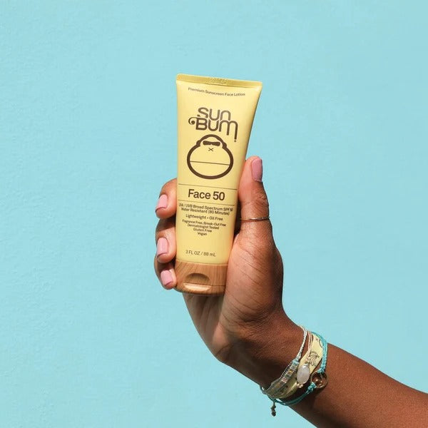 Hand holding a tube of Sun Bum Face SPF 50 sunscreen against a light blue background